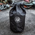 Load image into Gallery viewer, En person står på steiner ved vannet og holder en svart NEST-tørrbag med logo på.
